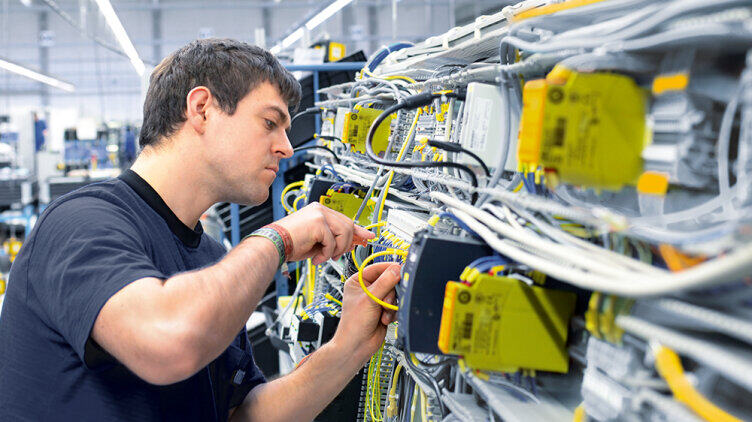 A person inspects and wires cables in a control cabinet containing safety and automation technology components